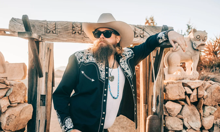 Robert Jon stands before a rustic wooden and stone archway, wearing glasses and a cowboy hat.