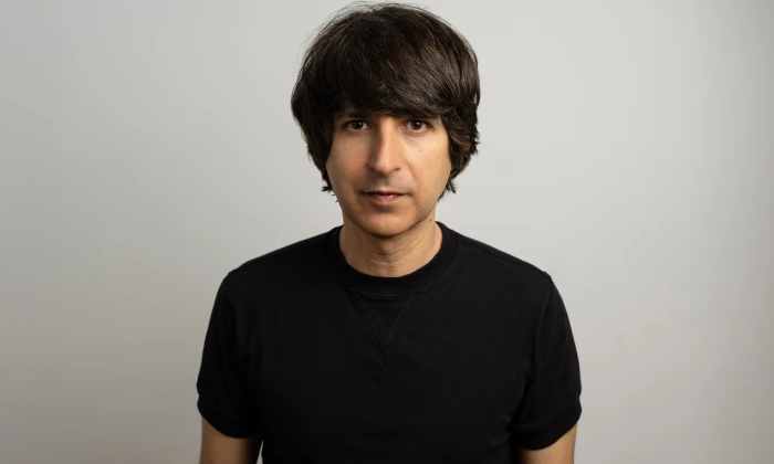 Demetri Martin dons a black shirt and poses in front of a gray backdrop.