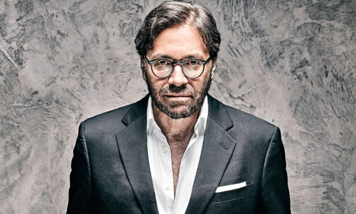 Al Di Meola dons a grey suit while posing against a textured backdrop.
