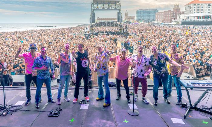 The Beach Boys smile and pose on stage with a lively concert crowd behind them.