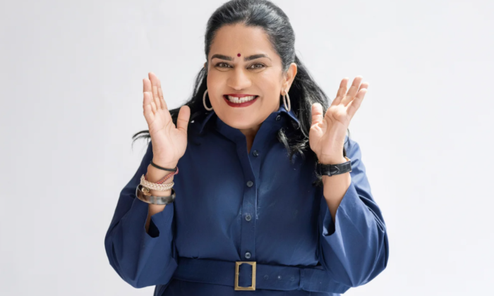Zarna Garg stands before a white backdrop, beaming in a blue dress.