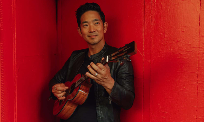Jake Shimabukuro smiles and strums his ukulele in front of a red backdrop.