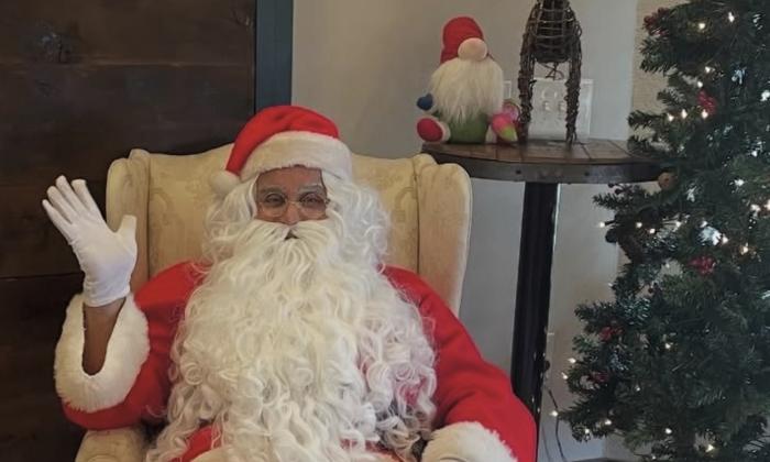 Santa waving from a comfortable chair, next to a Christmas Tree