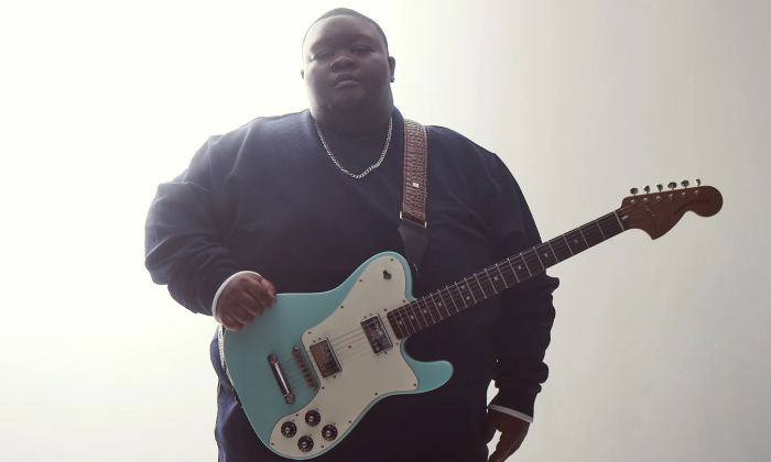 Christone "Kingfish" Ingram clutches his guitar and poses in front of a white backdrop.