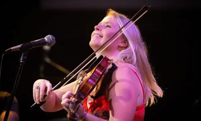 A smiling Charlotte Morris with her fiddle, on stage at a microphone