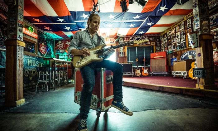 Chris Duarte sits on a barstool and strums his guitar.