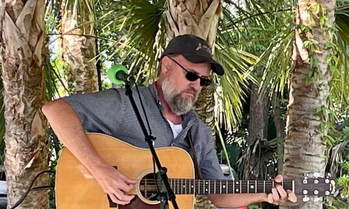songwriter Joey Kerr with his guitar, onstage, with palm trees behind.