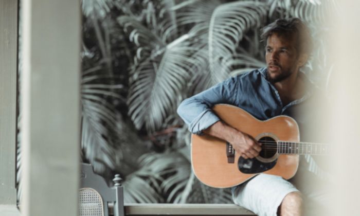 Mishka sits in a chair and strums his guitar.