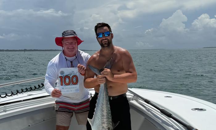 Two men on the foredeck of a fishing holding a kingfish and a sign with their boat's number