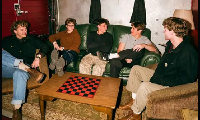 Bandmates from The Band Solstice sit on couches and pose in front of a brown table with a checkerboard. 