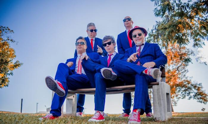 The Blues Beatles, dressed in blue suits and matching ties, sit together on a small bench for a posed shot.