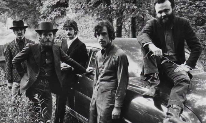 The Band’s members, dressed in dark attire, cluster together beside a vintage car.