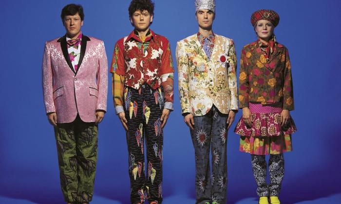 The Talking Heads wear colorful clothing and pose in front of a blue backdrop.