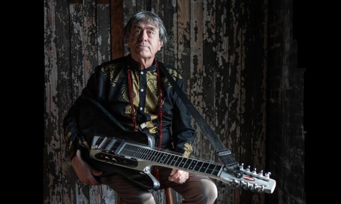 Arvid Smith sitting agaisnt a dark barnwood wall, holding a stringed instrument