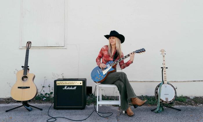 Lexi Long, before or after a show, sitting against a white wall, with two guitars, a banjo, and an amp