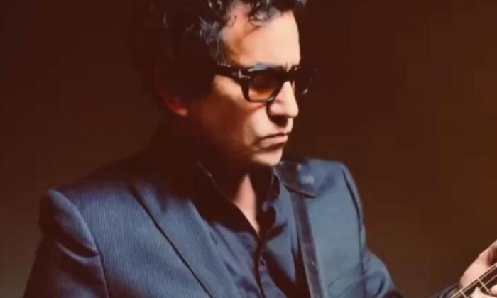 A.J. Croce wears dark sunglasses and poses in front of a dark brown backdrop.