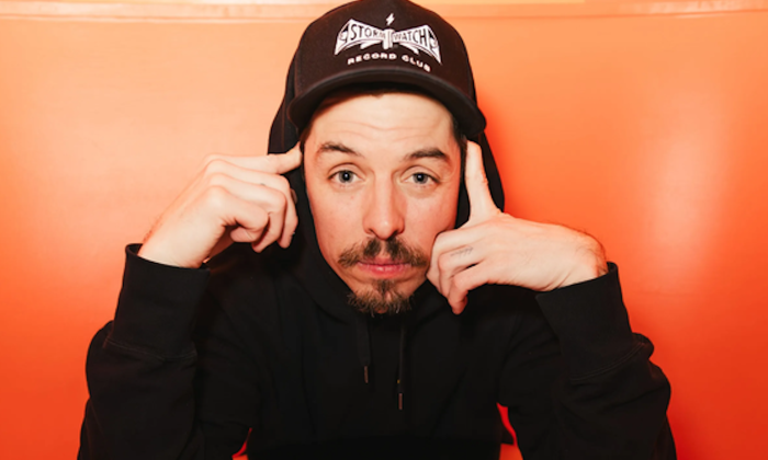 Grieves dons a black hoodie and poses in front of an orange backdrop.