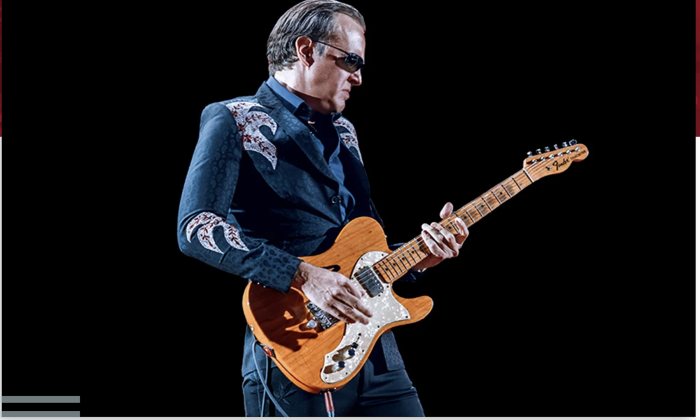 Joe Bonamassa strums his guitar in front of a black backdrop.