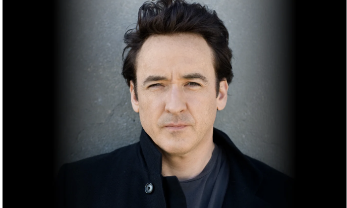 John Cusack wears black clothing and poses in front of a black and grey backdrop.