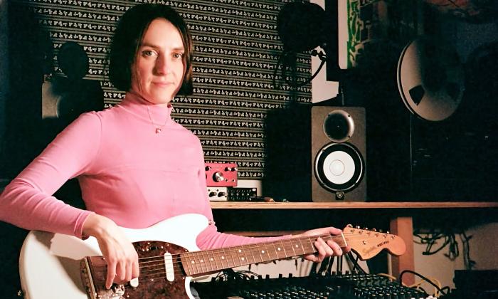 The leader of the band Slippers, wearing a pink turtleneck, in a music studio playing electric guitar