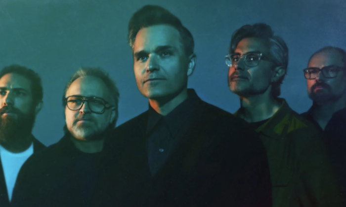 Bandmates from Death Cab for Cutie wear black clothing and pose in front of a dark blue backdrop.