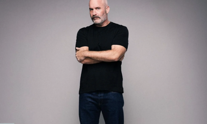Tom Segura dons black clothing and poses in front of a grey backdrop.
