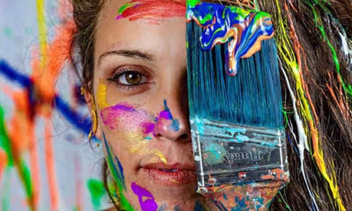 A woman with paint on her face, holding a housepainting brush up to cover her left eye