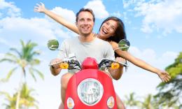 A young white couple rides a red moped towards the camera on a sunny day. They’re both smiling widely and the woman spreads her arms in glee