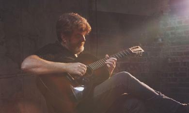 A musician playing guitar in a darkened room, with hazy sun shining on him