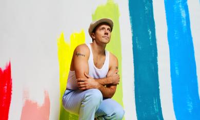 Jason Mraz, in cap, t-shirt, and jeans, in front of a rainbow-striped backdrop