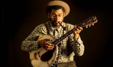American Songster Dom Flemons playing banjo against a dark background
