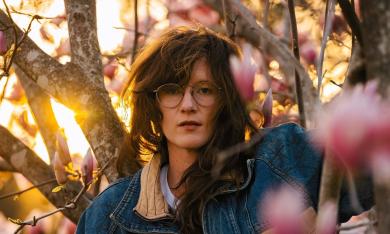 SG Goodman (a white woman with glasses and long tousled hair wearing a jean jacket) stares at the camera from the branches of a flowering tree. It is sunset