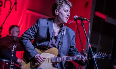 Gabe Stillman sings on a stage with a red backdrop. 
