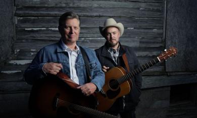The Gibson Brothers pose with their guitars in front of a wooden background. 