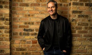 Pat McGann smiles and poses in a leather jacket in front of a brown brick wall. 