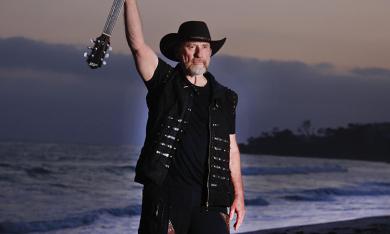 Colin Hay poses in black clothing while holding his guitar. 