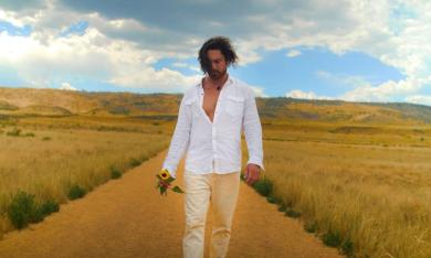 Will Evans wears a white top and holds a wilted sunflower while walking down a dirt road. 