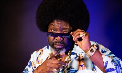 Afroman clutches his black sunglasses in front of a royal purple backdrop. 