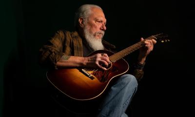 Jorman Kaukonen in jeans and a plaid shirt, strumming an acoustic guitar in a darkened stage.