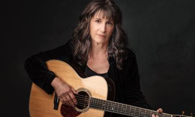 Karla Bonoff poses with her brown guitar in front of a black background. 