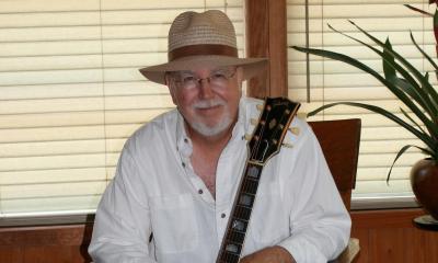 Larry Mangum, a singer and songwriter, sits in front of a window with his guitar
