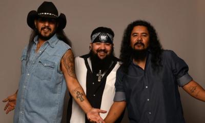 Bandmates from The Lonely Boys smile and pose in front of a brown backdrop.