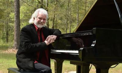 Chuck Leavell smiles and leans on a black piano.