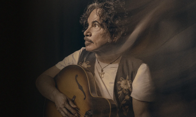 John Oates strums his guitar in front of a dark brown backdrop.