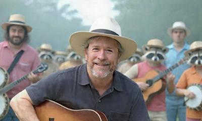 Songwriter Kevin Edwards with his guitar, outside, backed by banjo playing racoons and others