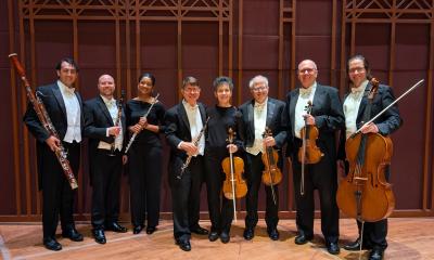 Eight musicians in a winds and strings ensemble, standing in a room with majestic wood paneling