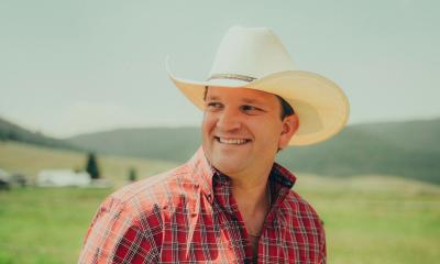 songwriter David Adam Byrnes in white cowboy hat, grinning, with hills shown behind him