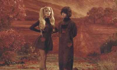 Domi and JD Beck sport burnt-orange outfits, posing against a warm, autumnal backdrop.