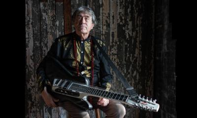 Arvid Smith sitting agaisnt a dark barnwood wall, holding a stringed instrument