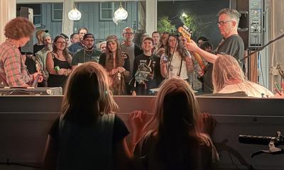 A band trio, playing outside to an audience, while two little girls watch from the inside through a window